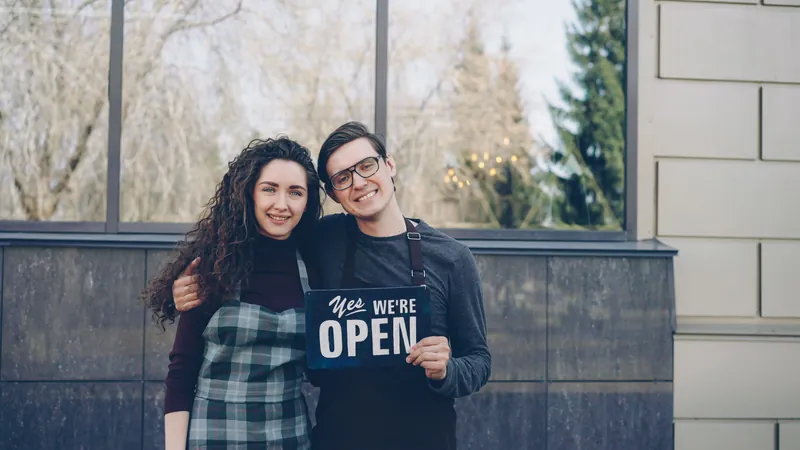 Two small business owners holding an open sign outside their storefront